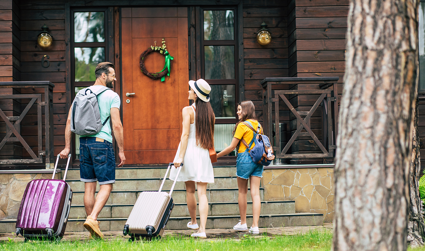 Summer joy. A photo from behind of a happy travelling family with suitcases and backpacks coming to the modern wooden hotel, father and mother looking at each other.