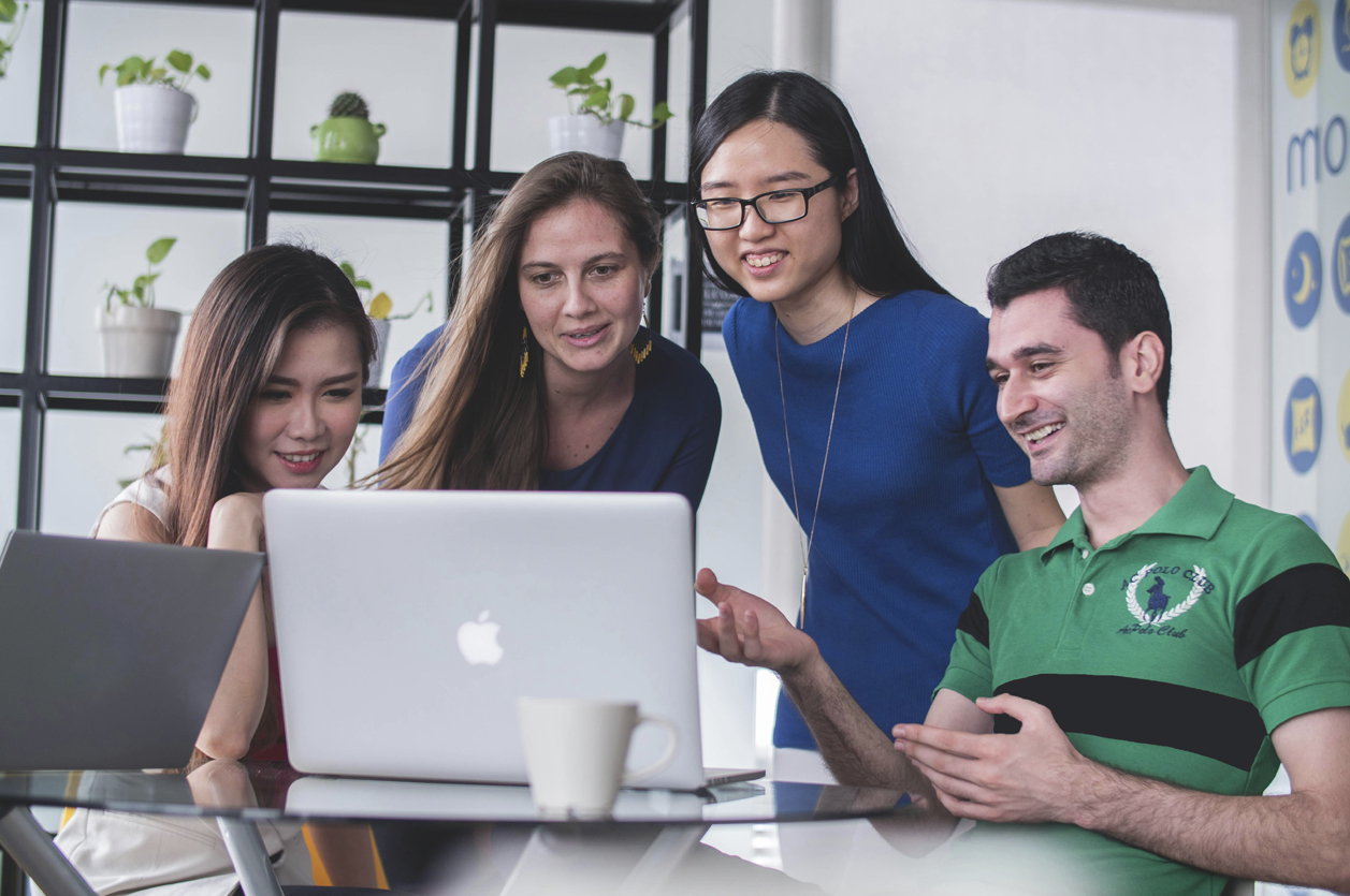 A group of young multiethnic office colleagues gather around a laptop computer. Happy, engaged team members review something on the computer screen.