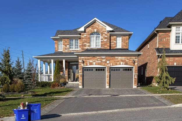 Exterior of large two-story brick home with two-car garage. Large blue recycling bins at the roadside and deep blue skies all around.