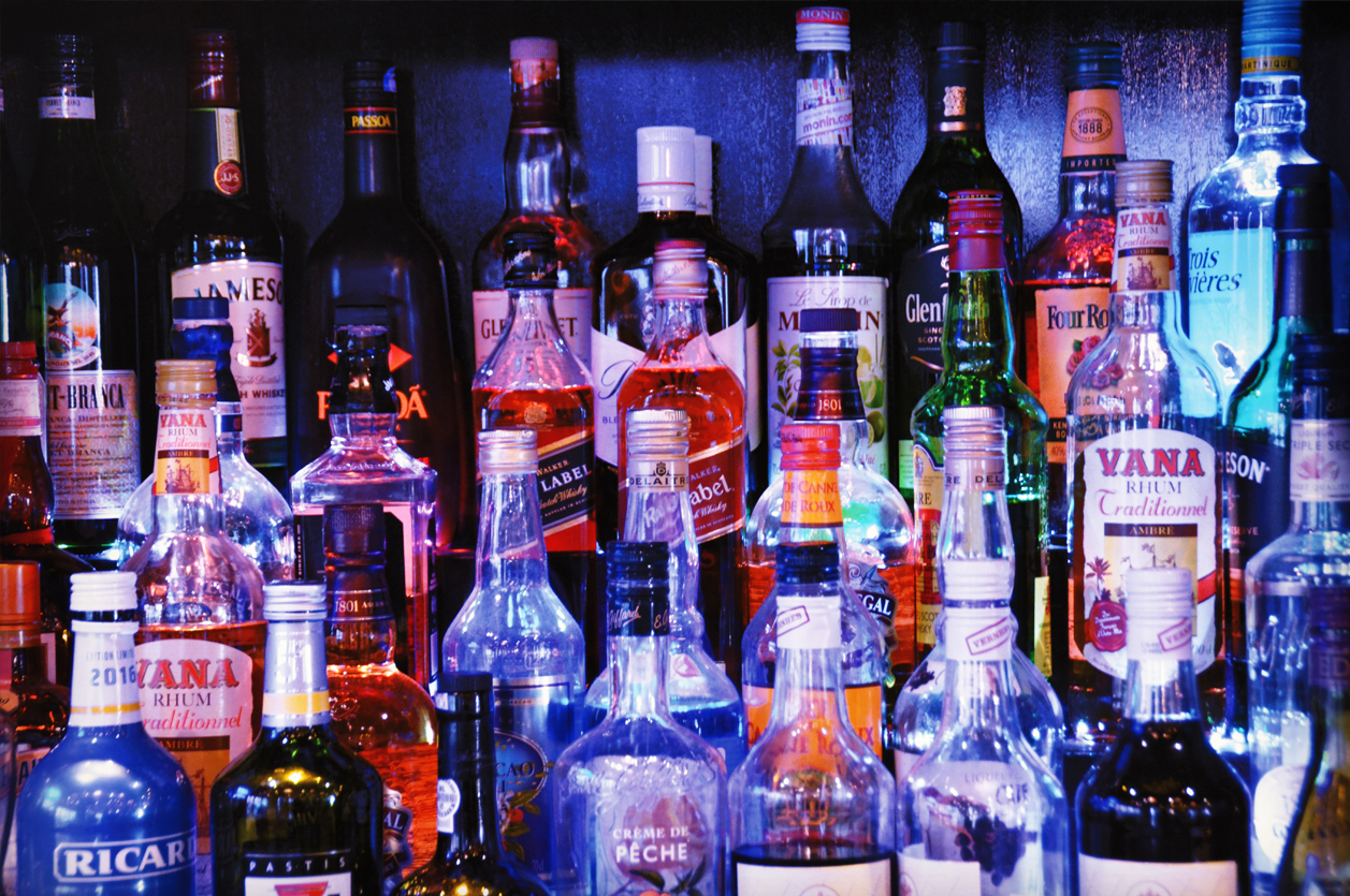 A dramatically-lit bar display of dozens of different liquor bottles, many partially empty. Glass bottles of rum and whiskey shine, reflecting the neon bar lights.