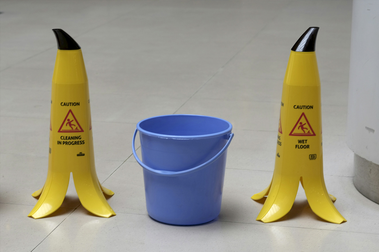 Two yellow 'wet floor' safety signs beside a bucket collecting water leaking from the ceiling. The signs are in the shape of banana peels to attract attention and warn against slipping.