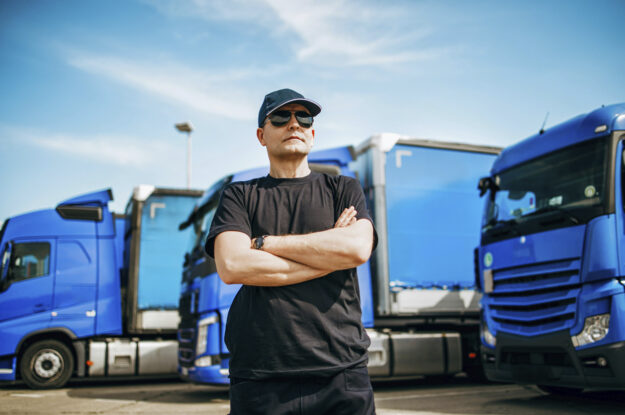 Truck driver in sunglasses wearing all black stands defensively with his arms crossed in a truck yard with several tractor trailer trucks in the background.
