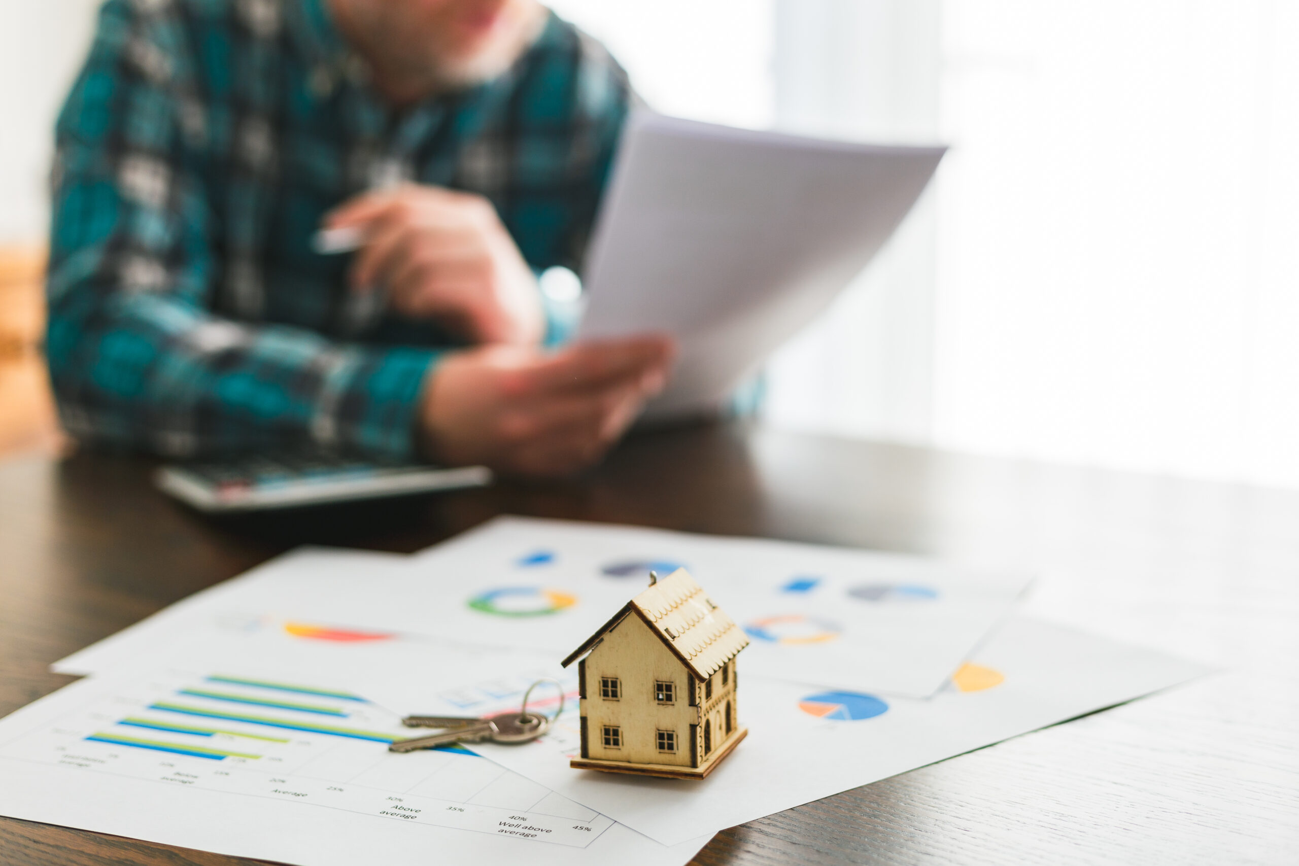 Signing a house sale agreement A man looking at documents pertaining to a house sale agreement. Focus on a model house on top of documents in the foreground, while the man - sitting at a table and studying the agreement papers - is defocused beyond.