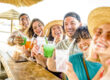 Happy multi ethnic friends cheering and drinking coloured cocktails at a beachfront tiki bar, celebrating summer.