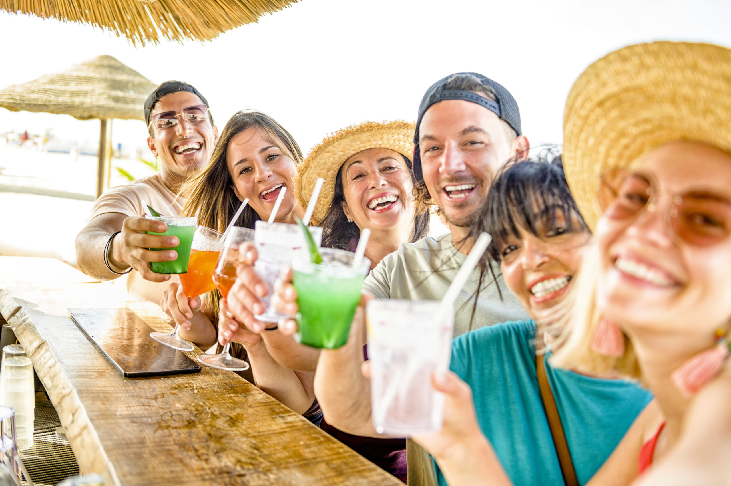 Happy multi ethnic friends cheering and drinking coloured cocktails in kiosk at beach – Young millennials and senior people having fun at summer weekend – Selfie lifestyle and party concept Happy multi ethnic friends cheering and drinking coloured cocktails at a beachfront tiki bar, celebrating summer.