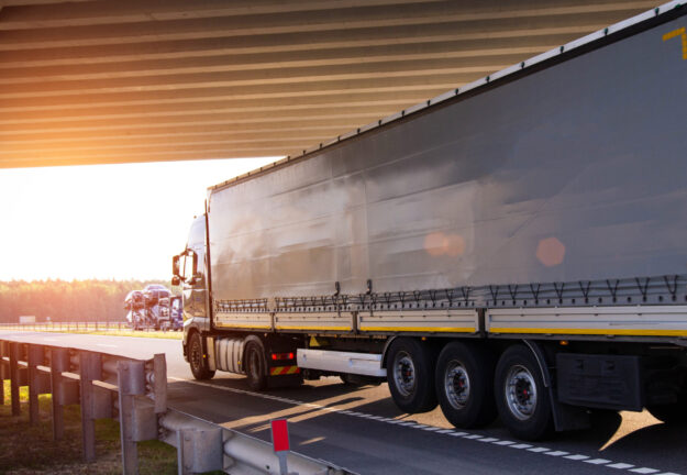 A truck with a tilt semi-trailer carries cargo under a bridge against the backdrop of sunset, copy space for text. Logistics A truck with a tilt semi-trailer carries cargo under a bridge against the backdrop of sunset
